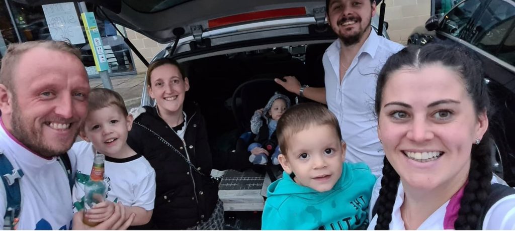 Laura and Mark with their godsons Lucas, Jacob and Isaac (from left to right), and Naomi and Nathan are posing next to a car. They are all smiling at the camera.
