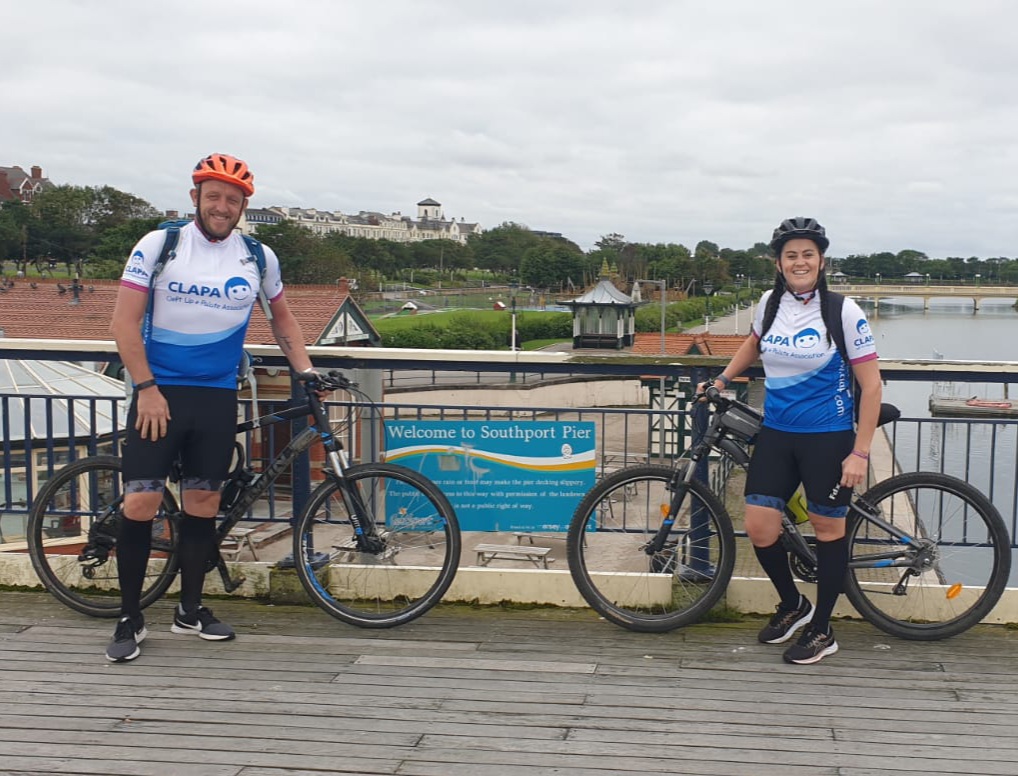 Laura and Mark are standing on a bridge by a sign that says 'Welcome to Southport Pier'. They are both standing next to their bikes and smiling at the camera. 