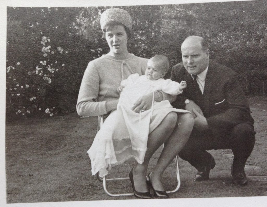 A black and white photo of Helen, wearing a smart dress, heels and a hat, sitting on a chair holding baby Sean who wears a long white Christening robe. A man dressed in a suit crouches next to the chair.