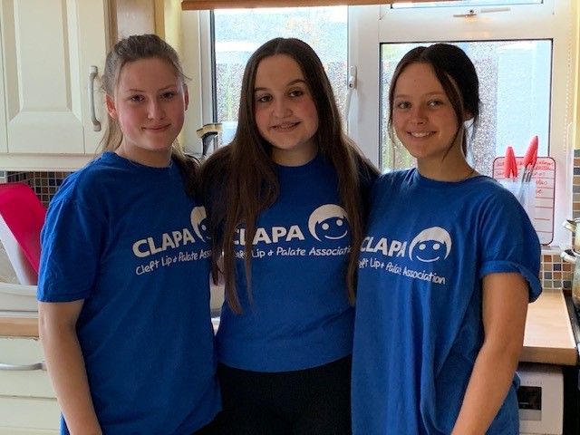 Three girls stand next to each other and wear blue CLAPA t shirts while smiling at the camera.