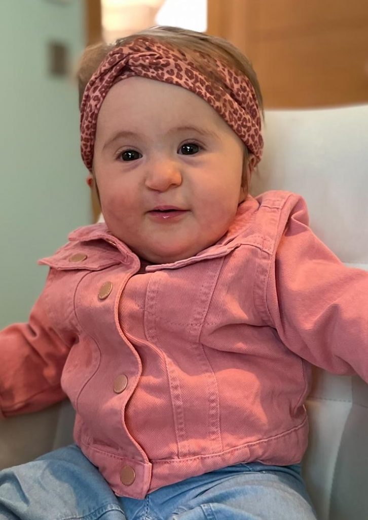 A young girl sits upright on a sofa wearing a pink jacket and pink headband. She is looking at the camera and smiling gently.