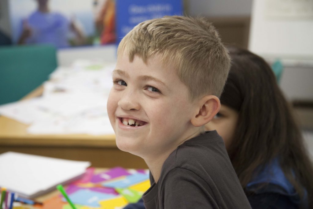 a boy smiles widely at the camera with pieces of paper and pens out of focus in the background behind him