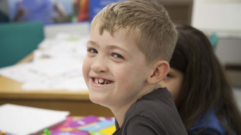 a boy smiles widely at the camera with pieces of paper and pens out of focus in the background behind him