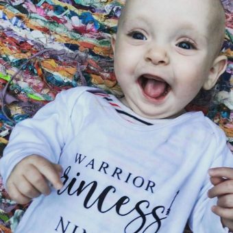 a baby is smiling widely wearing a white top with text reading 'warrior princess'