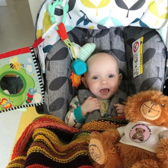 A baby sits in a grey baby seat surrounded by toys and smiling widely at the camera