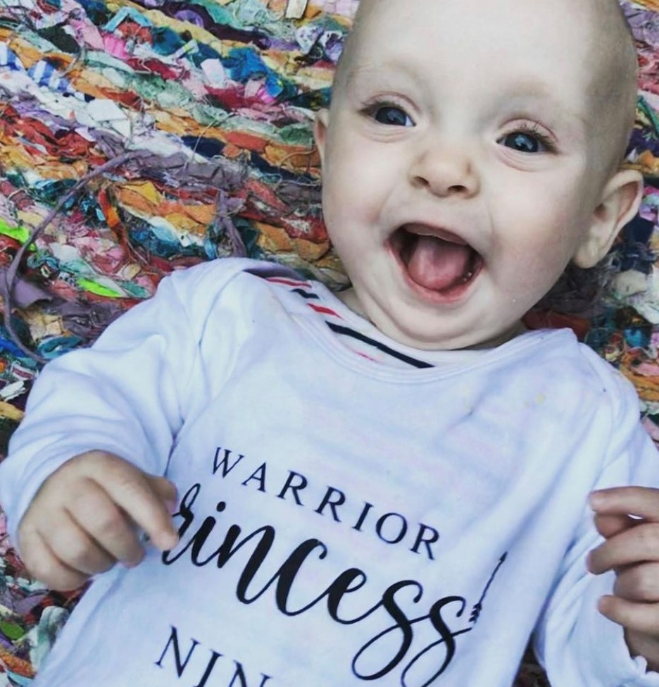 a baby is smiling widely wearing a white top with text reading 'warrior princess'