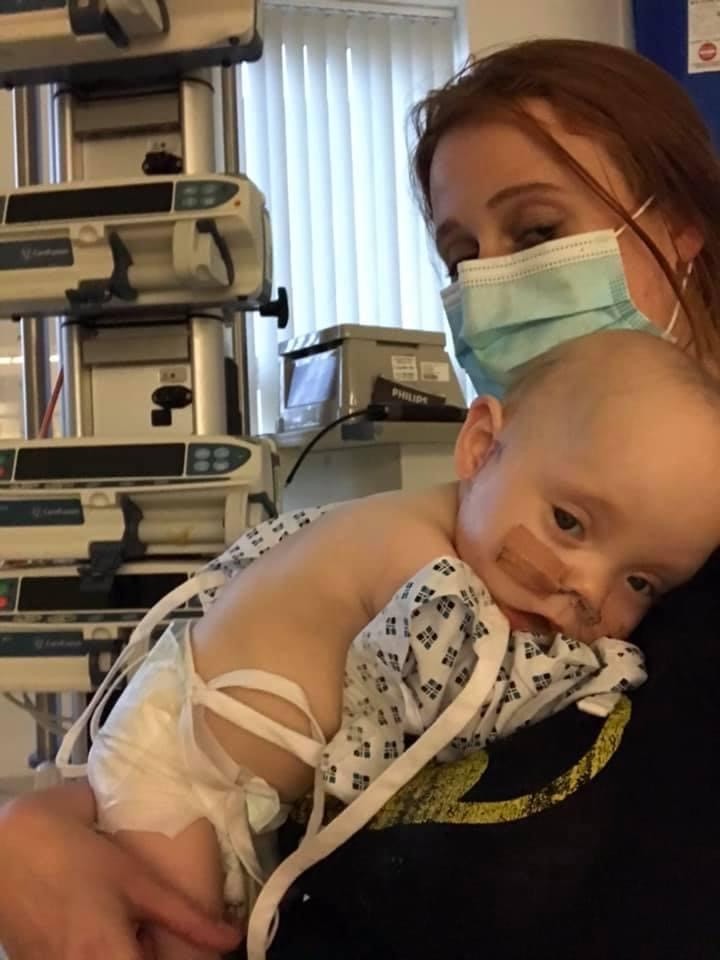 A woman wearing a mask holds a baby with a feeding tube who is wearing a hospital gown