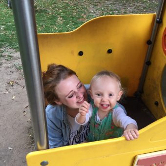 A woman holds a smiling baby while crouched down in a playground