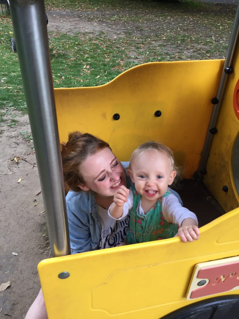 A woman holds a smiling baby while crouched down in a playground