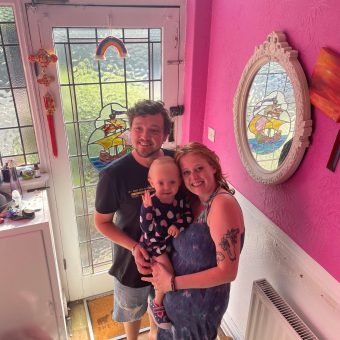 A man and woman hold a toddler, and all are smiling at the camera in a hallway