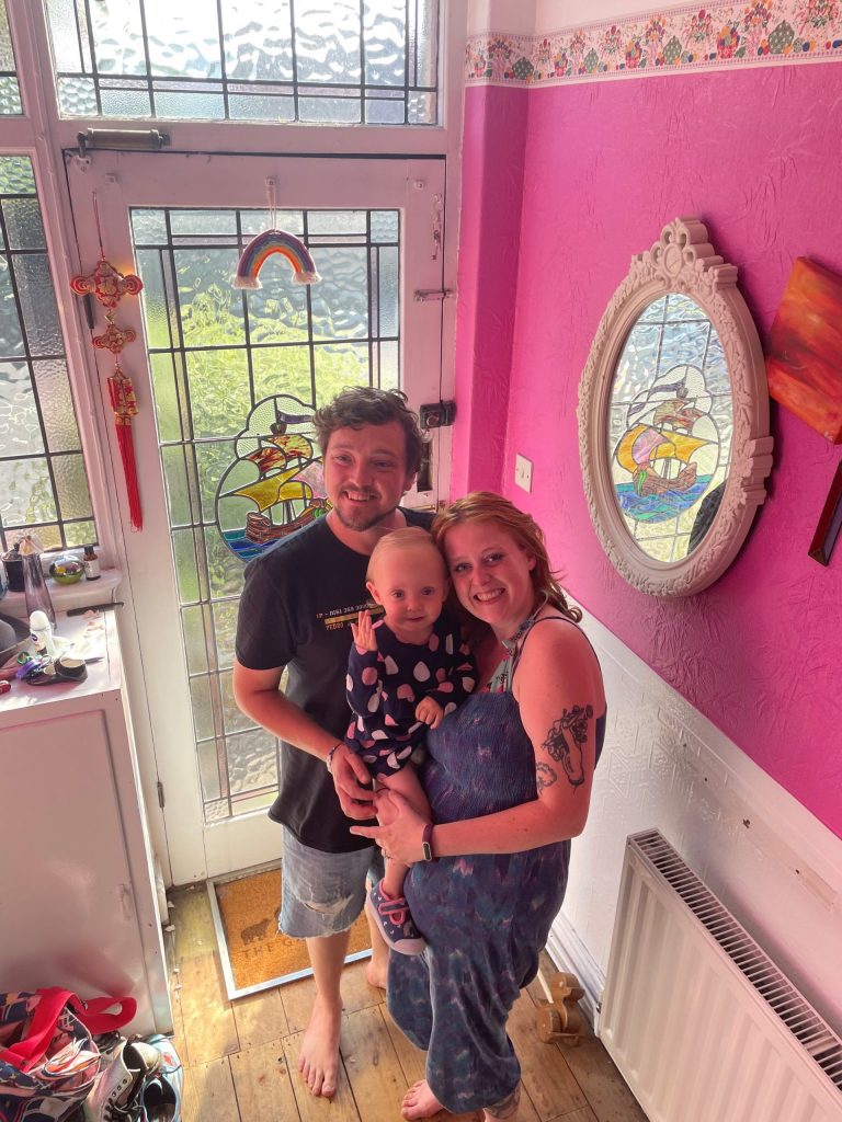 A man and woman hold a toddler, and all are smiling at the camera in a hallway