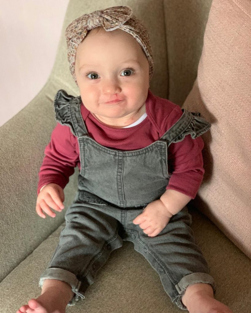 A baby with a faint scar by her lip is sitting up on a chair wearing dungarees and a headband