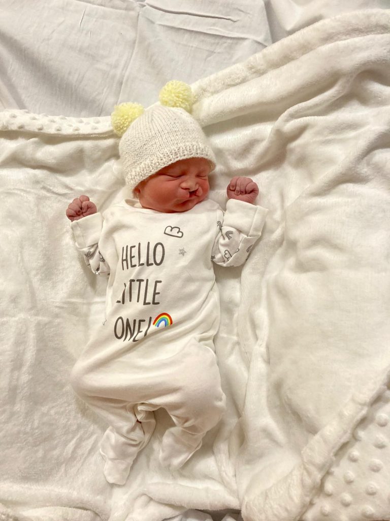 A newborn baby with a cleft lip is lying on a white sheet wearing a white babygrow and hat