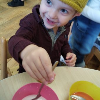 a young boy is holding a spoon upright in a milky bowl, looking away from the camera and wearing a yellow hat