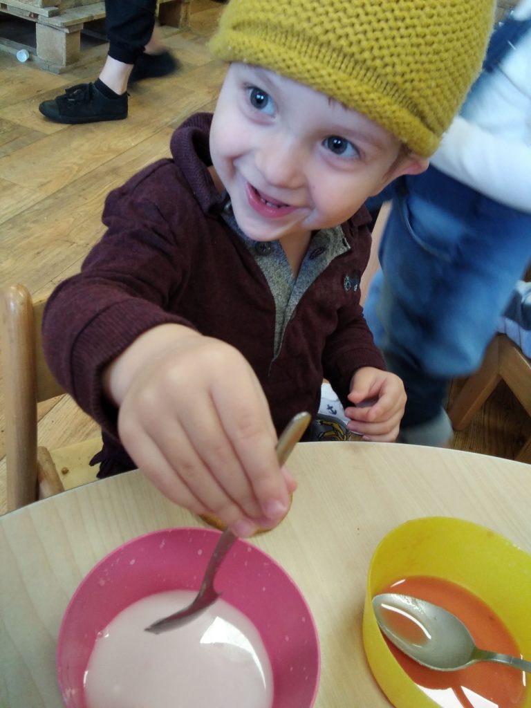 a young boy is holding a spoon upright in a milky bowl, looking away from the camera and wearing a yellow hat