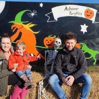 a woman holds a young child and sits on a bale of straw next to a man