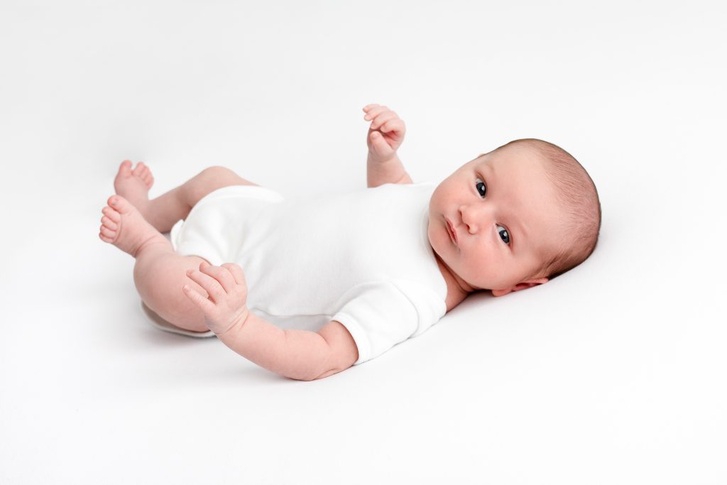 A white background with a baby looking at the camera, wearing a white babygrow