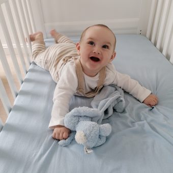 A baby lying down on their stomach in a coat, smiling and looking up. They are wearing beige dungarees and are next to a blue toy