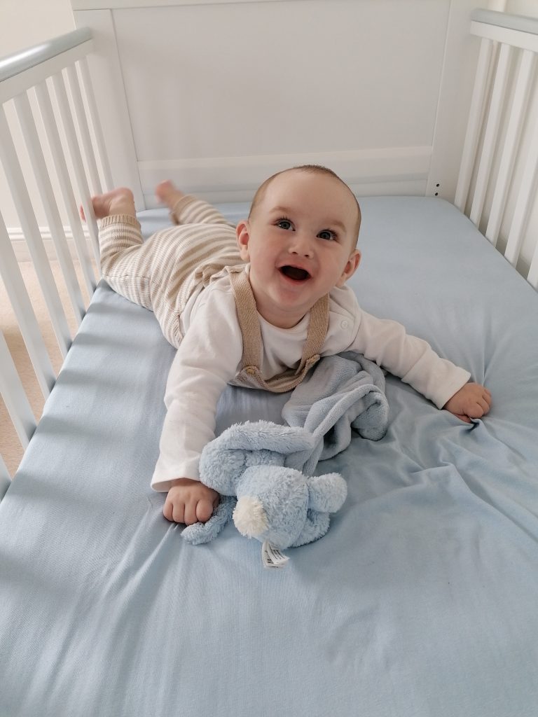 A baby lying down on their stomach in a coat, smiling and looking up. They are wearing beige dungarees and are next to a blue toy