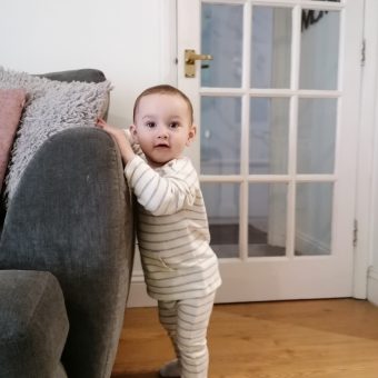 A toddler stands up, looking at the camera and leaning on the arm of a sofa with a door in the background