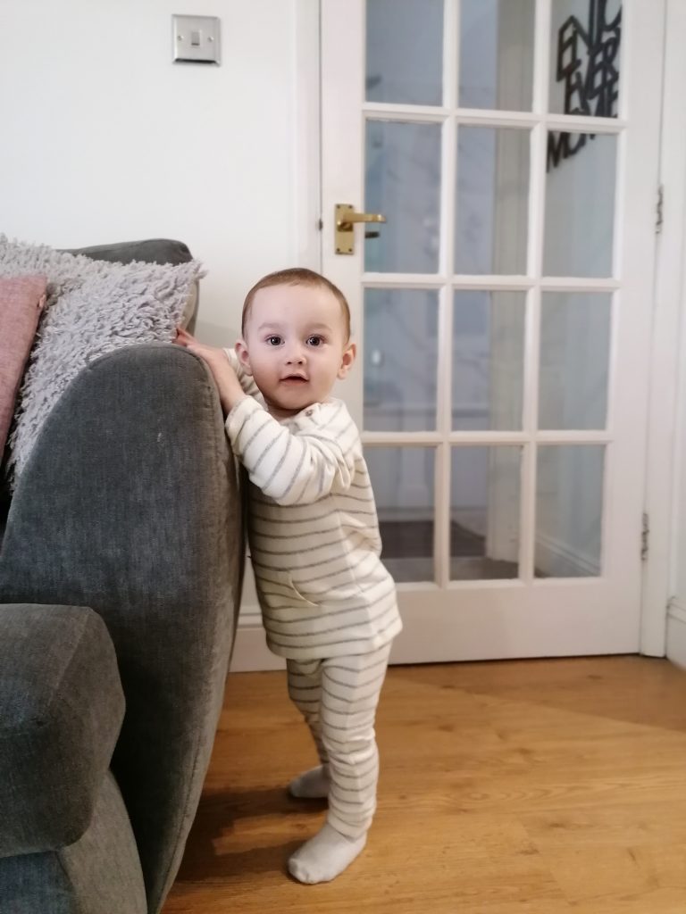 A toddler stands up, looking at the camera and leaning on the arm of a sofa with a door in the background