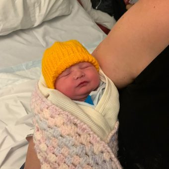 A baby being held, wrapped in a blanket and wearing a yellow hat