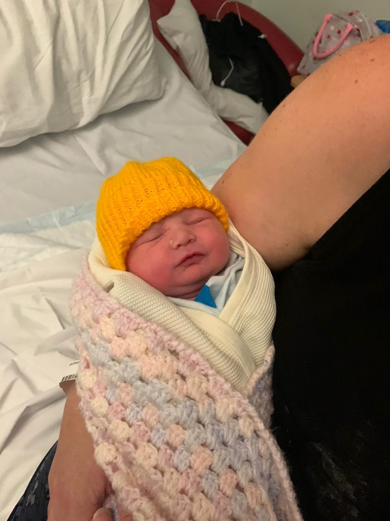A baby being held, wrapped in a blanket and wearing a yellow hat