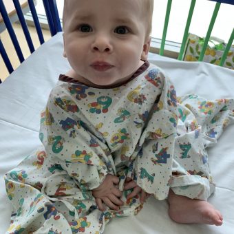 A baby wearing a hospital gown sits up and looks at the camera