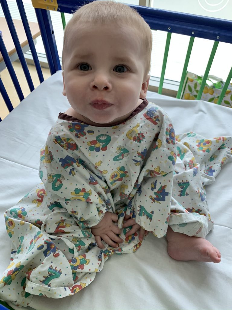 A baby wearing a hospital gown sits up and looks at the camera