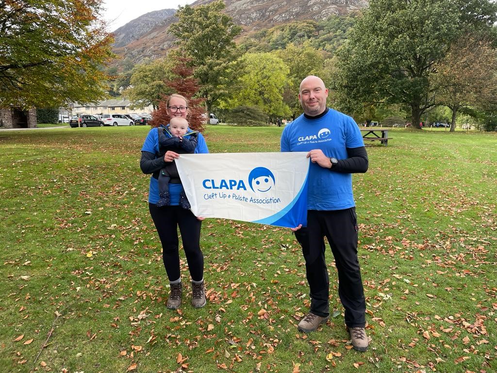 A woman and man wearing blue CLAPA t shirts hold a CLAPA banner. The woman is holding a baby in a baby carrier on her chest.