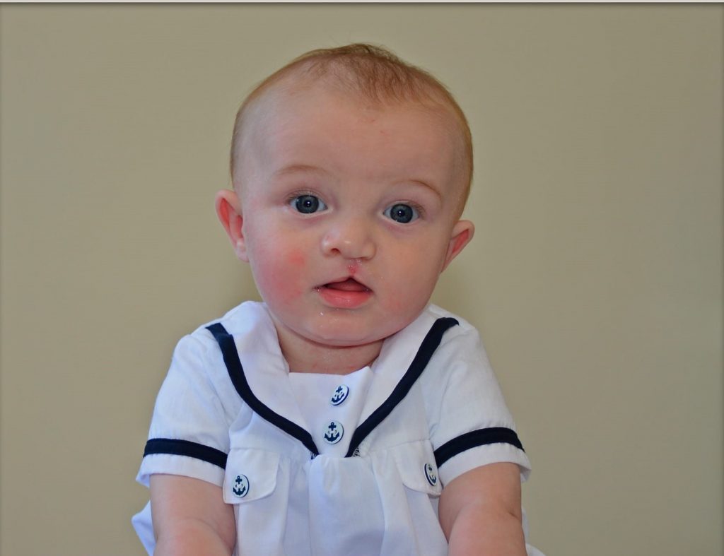 A baby in a white sailor shirt looks at the camera and sits on a blanket patterned with elephants