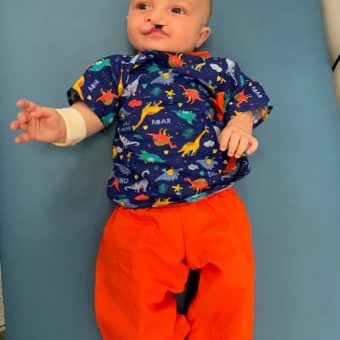 A baby in a dinosaur top and red trousers lying on a hospital bed