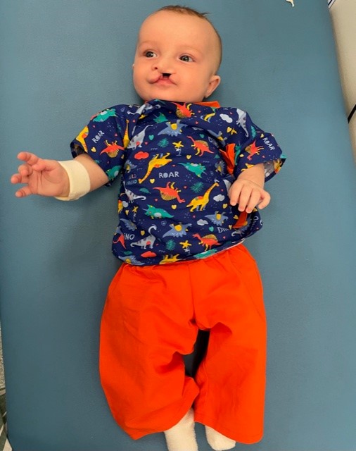 A baby in a dinosaur top and red trousers lying on a hospital bed