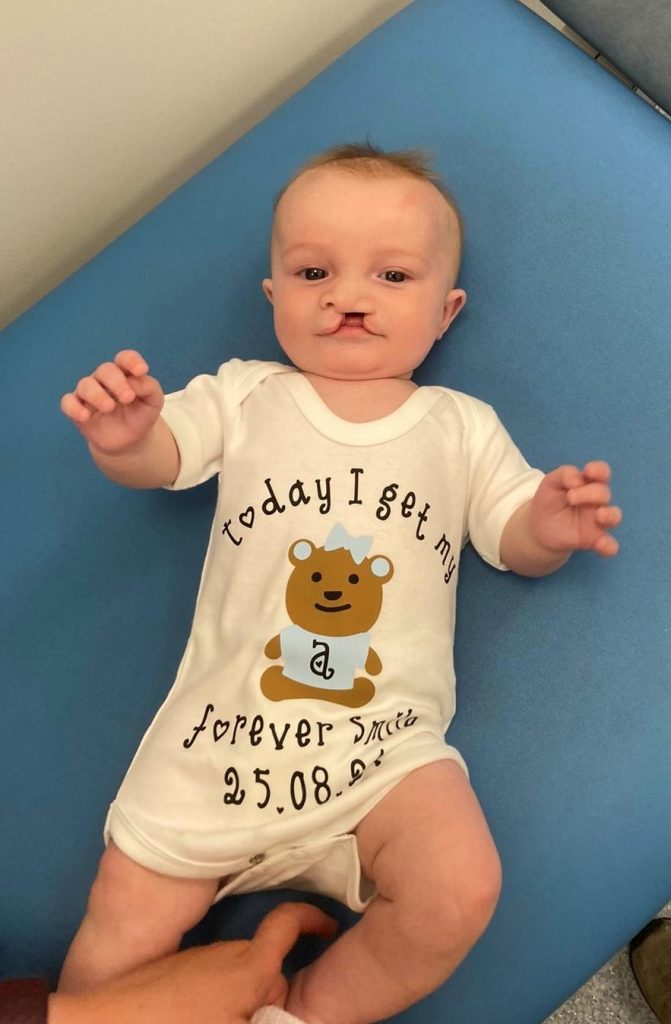 a baby lying on a hospital bed wearing a babygrow saying 'today I get my forever smile';
