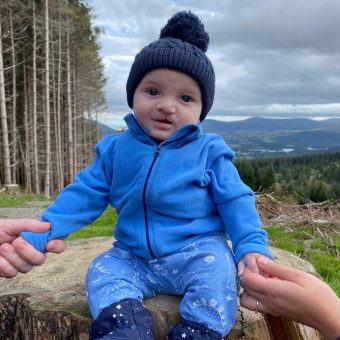 A baby in blue clothes and a dark blue hat sits on a tree stump with trees and mountains in the background behind