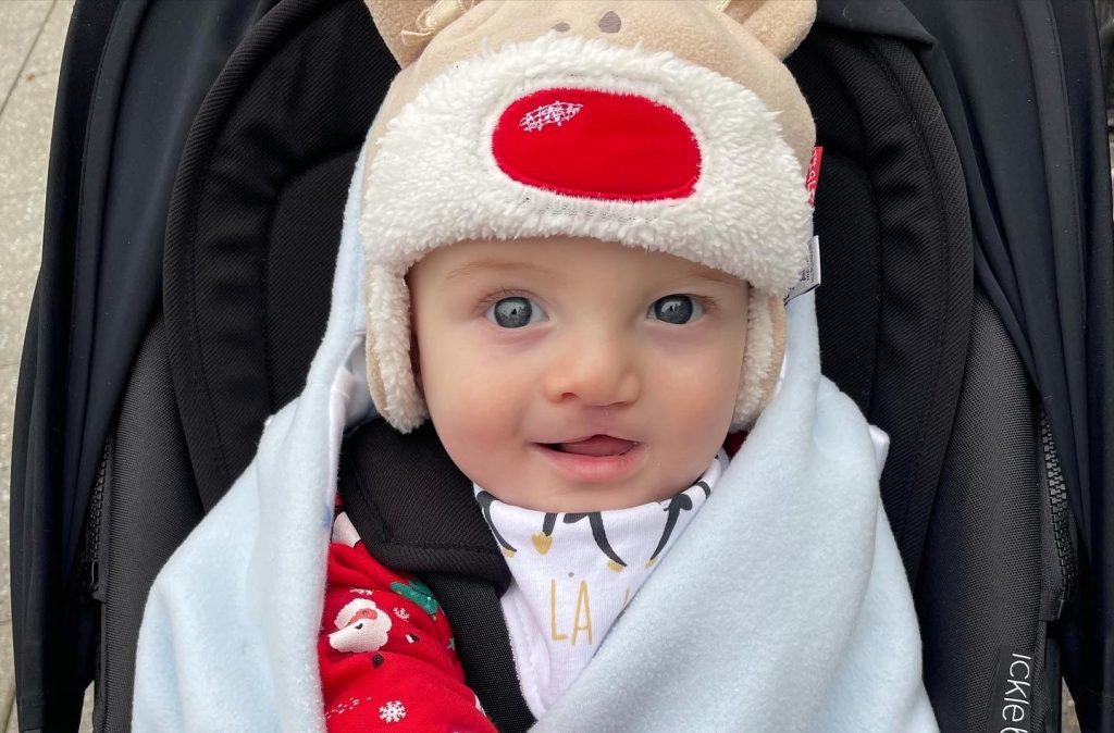 A baby wearing a reindeer hat and sitting in a pram, looking at the camera