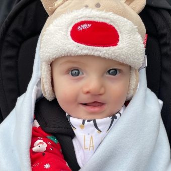 A baby wearing a reindeer hat and sitting in a pram, looking at the camera