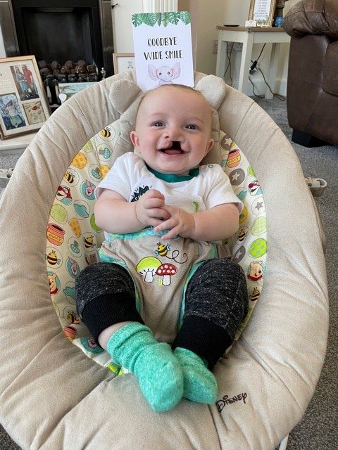 A baby in a baby seat with a sign saying 'goodbye wide smile'