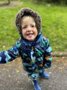 A young boy wearing a bue coat and wellies stood on a path by grass