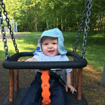 A baby at a park sat in a swin, wearing a light blue t-shirt and hat