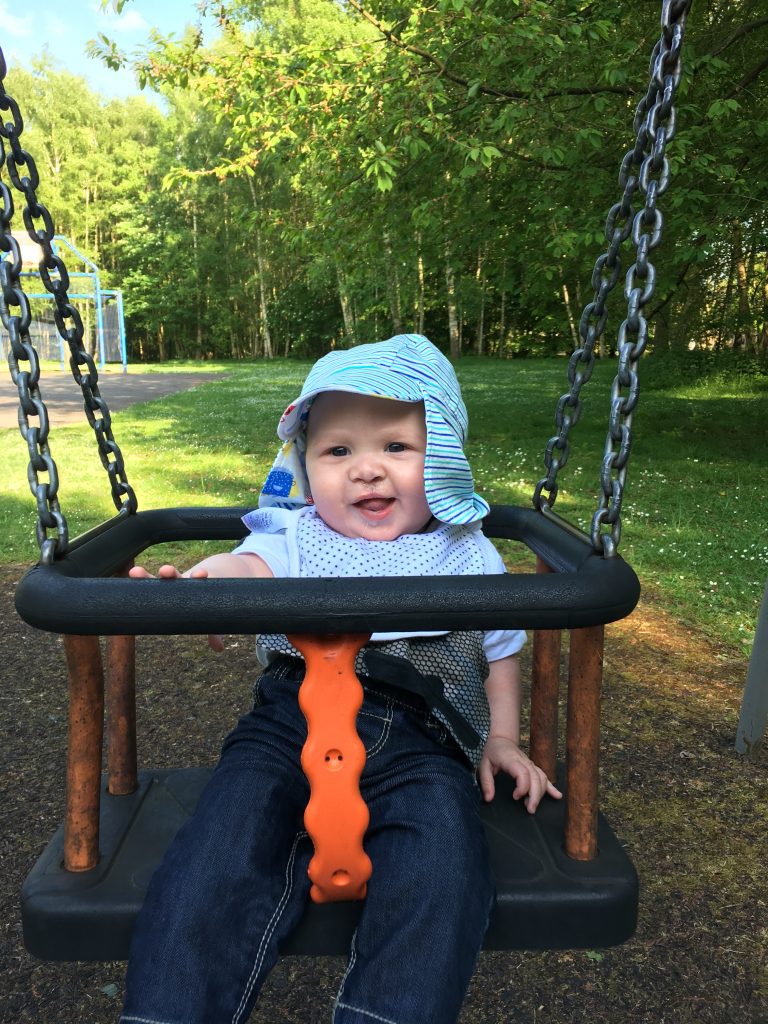 A baby at a park sat in a swin, wearing a light blue t-shirt and hat