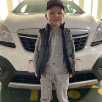 A young boy stood in front of a white car. hE WEARS A GREY TRAKSUIT AND A BLACK CAP.