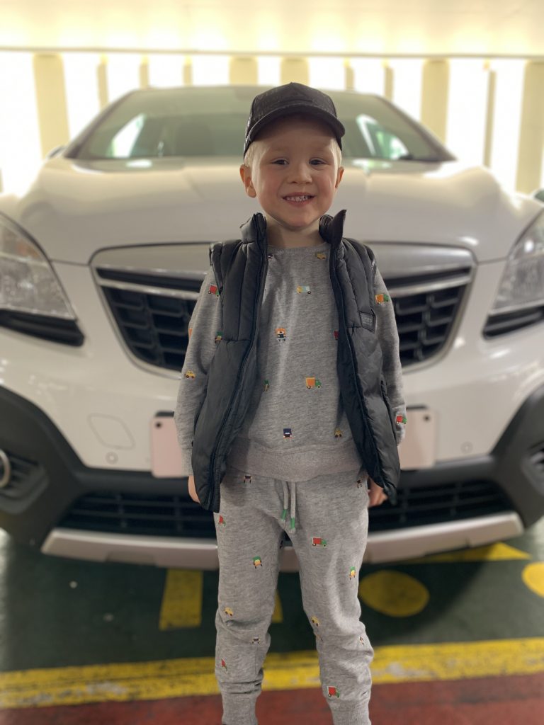 A young boy stood in front of a white car. hE WEARS A GREY TRAKSUIT AND A BLACK CAP.