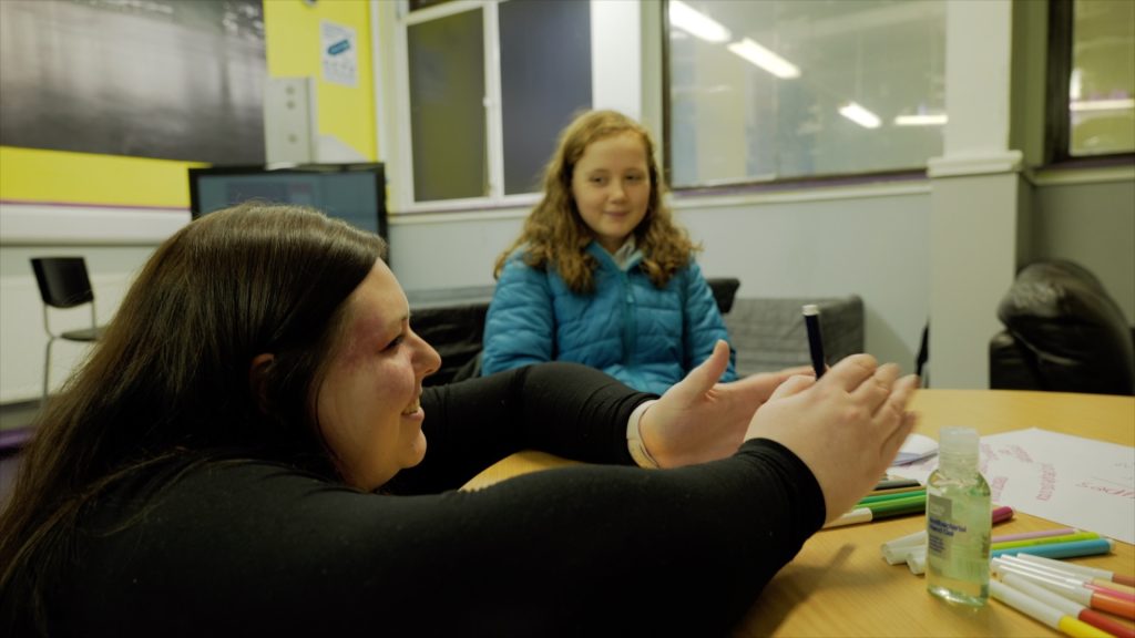 [Image ID: A female youth worker wearing a black top leaning on a desk, with a young girl wearing a blue coat. In the background is a classroom.]