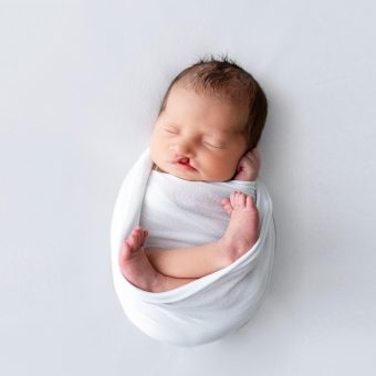 A newborn baby wrapped in white cloth against a grey background.