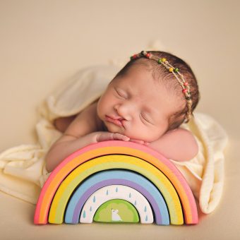 A small baby sleep on rainbow building blocks. She wears a cream dress against a cream background.