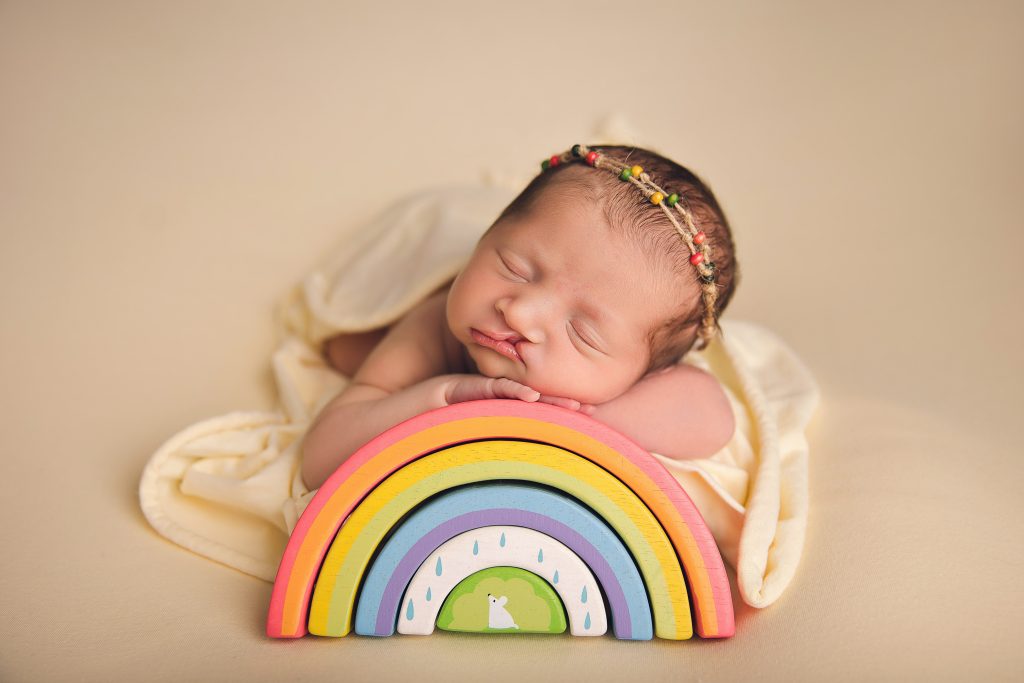 A baby asleep in a photo studio, leaning on rainbow blocks, wearing a cream dress with a cream background.