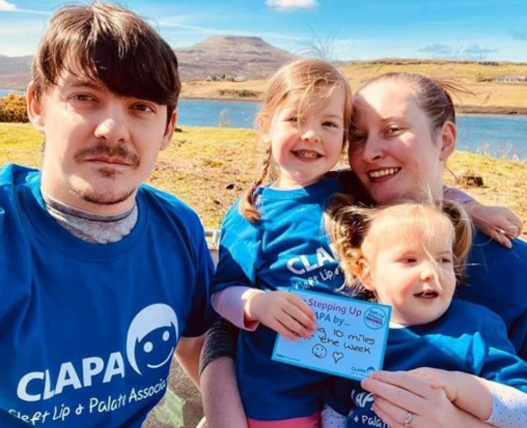 A family with two young children wearing CLAPA t shirts outside. A little girl is holding a 'Step Up' postcard which explains they're raising money for CLAPA by walking 10 miles during Awareness Week.