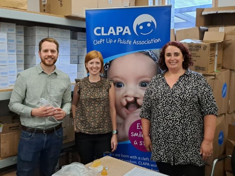 Photograph of a representative from the Croda Foundation and two CLAPA staff members standing in front of boxes of special feeding teats and a CLAPA banner.